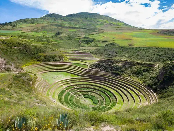 Terrazas agrícolas circulares de Moray en forma de anfiteatro verde, rodeadas de campos de cultivo bajo un cielo azul con nubes