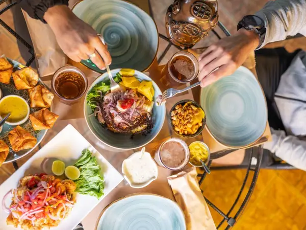 Mesa compartida con varios platos peruanos, salsas y bebidas, y manos de comensales sirviéndose