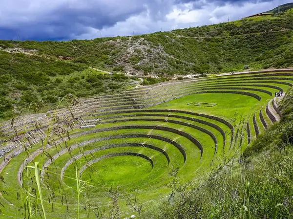 Andenes circulares de Moray cubiertos de pasto verde, con un grupo de visitantes en el sendero del borde y nubes de lluvia al fondo