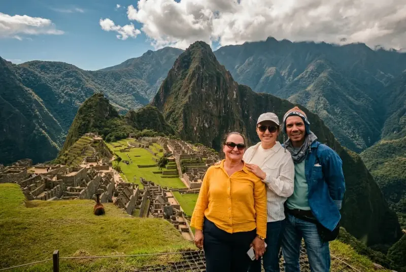 Tres viajeros sonrientes posando en Machu Picchu, con la ciudadela inca y la montaña Huayna Picchu detrás bajo un cielo con nubes