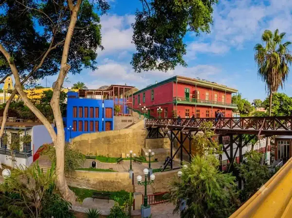 Puente de los Suspiros de madera en Barranco, entre árboles y casonas de colores rojo y azul, bajo un cielo despejado