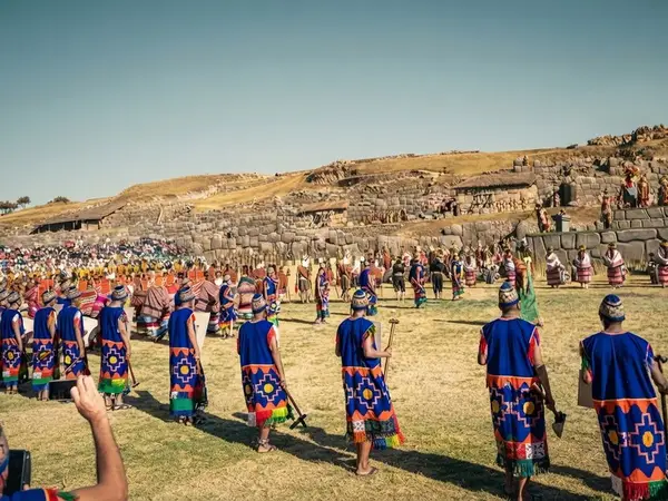 Danzantes con trajes azules y tocados avanzando en fila por la explanada de Sacsayhuamán durante el Inti Raymi, con público en las laderas
