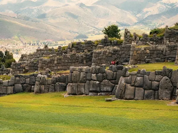 Muros incas de grandes bloques de piedra en Sacsayhuamán sobre una explanada de césped, con la ciudad de Cusco al fondo