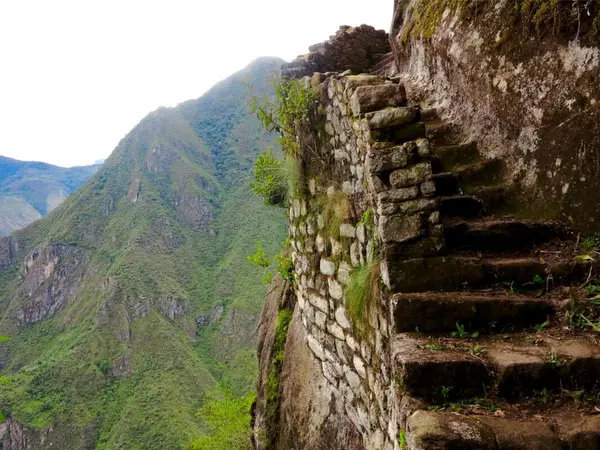 Escalinata inca de piedra en la subida a Huayna Picchu, pegada a la roca y al borde del precipicio, con montañas verdes al fondo