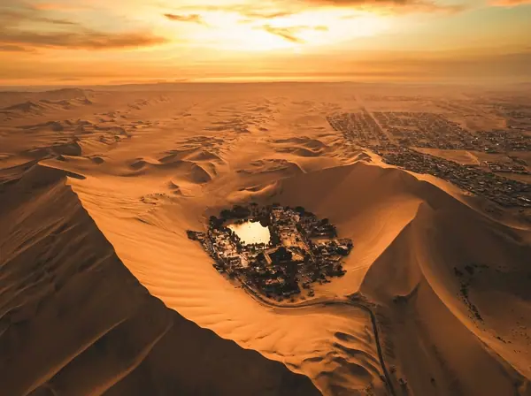 Aerial view of the Huacachina oasis surrounded by sand dunes at sunset, with the city of Ica in the distance