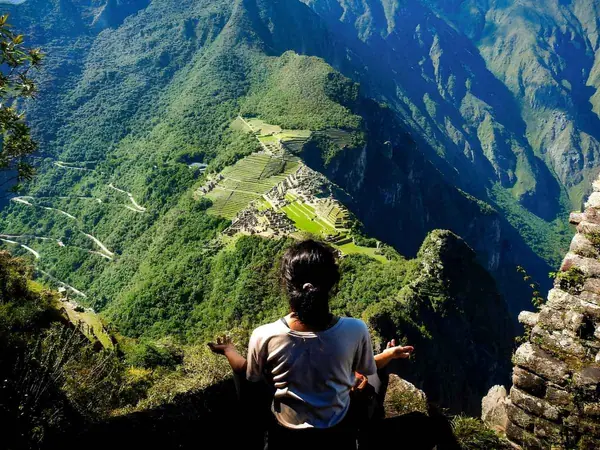 Traveller sitting on a mountain summit above Machu Picchu, with the citadel and the zigzag bus road far below