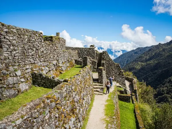 Hikers walking a stone path between the high walls of Sayacmarca, an Inca ruin on the Inca Trail, above green mountain slopes