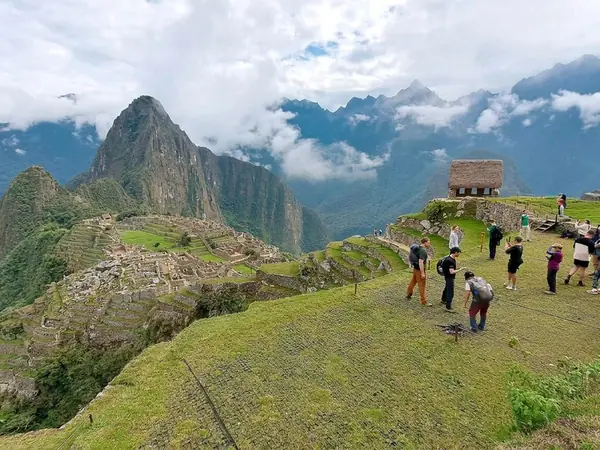 Visitors on the upper terraces of Machu Picchu near the thatched Guardian's House, with the city and Huayna Picchu in the distance