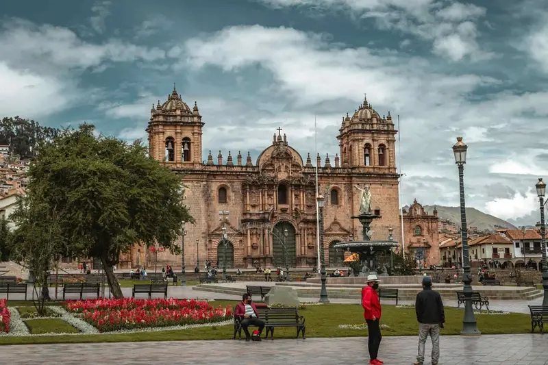 Cusco's main square with the stone cathedral and its two bell towers, flower beds and a fountain under a cloudy sky